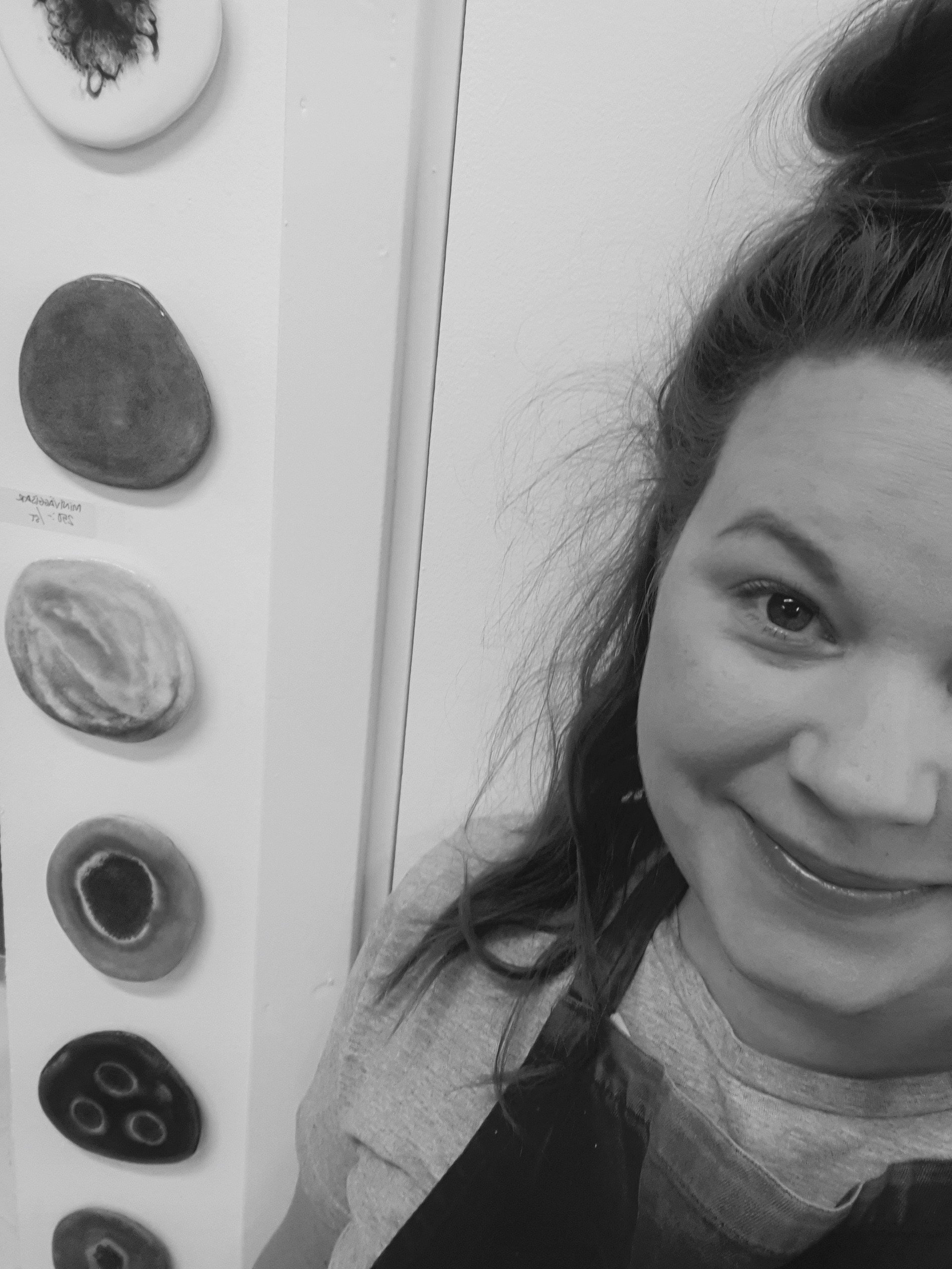 En glad kvinna med brunt hår i en hög tofs, delar av hennes ansikte syns, bakom henne finns en vägg med flera dekorativa stenar.