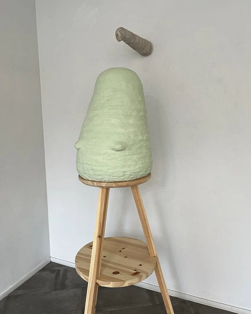 Ett konstverk som liknar en stor, grön, klot-formad figur med ansiktsdrag, placerad på en trästol. I bakgrunden syns en vägg och ett gardinliknande föremål.