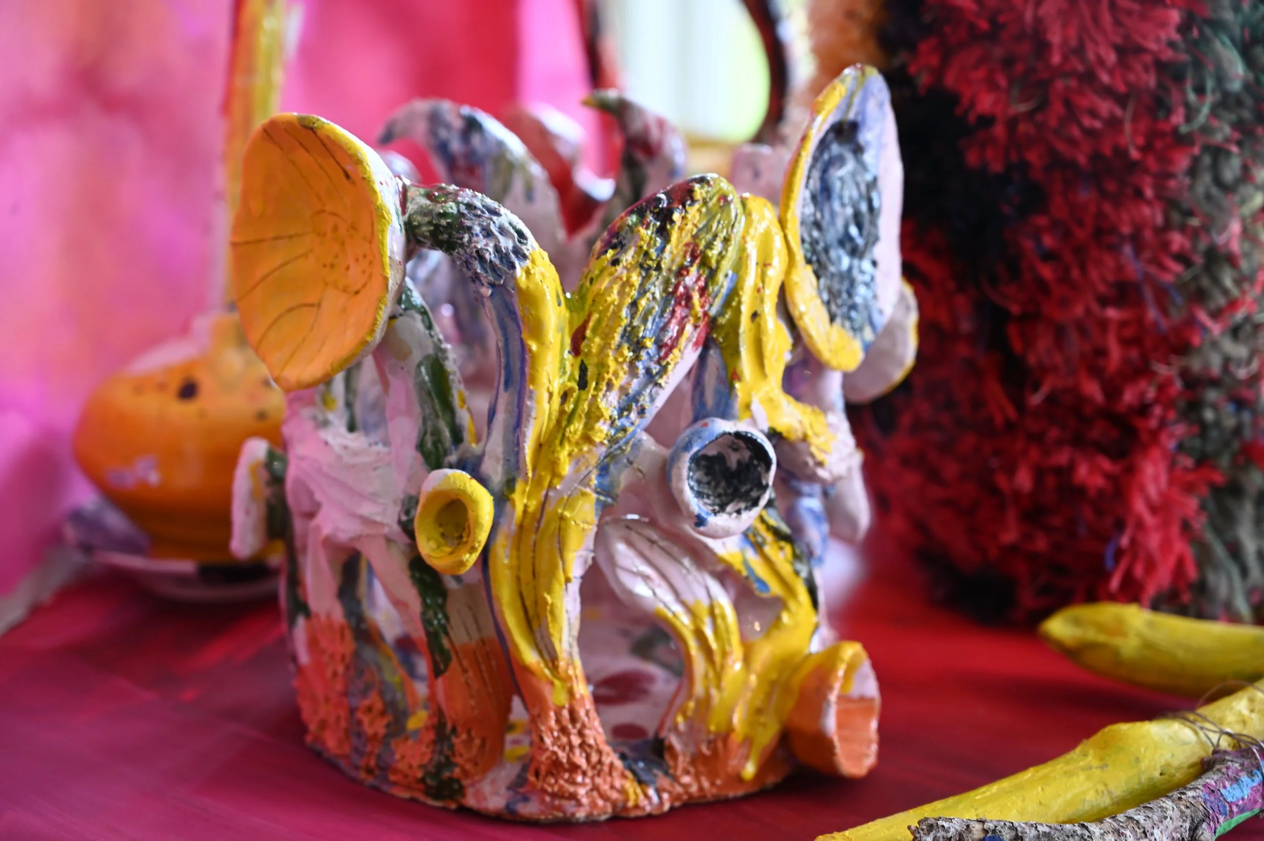 Färgglad keramikskulptur med oregelbundna former, målade i rosa, orange, gul, blå, grön och vit, placerad på ett rosa underlag med bakgrund i rosa och grönt.