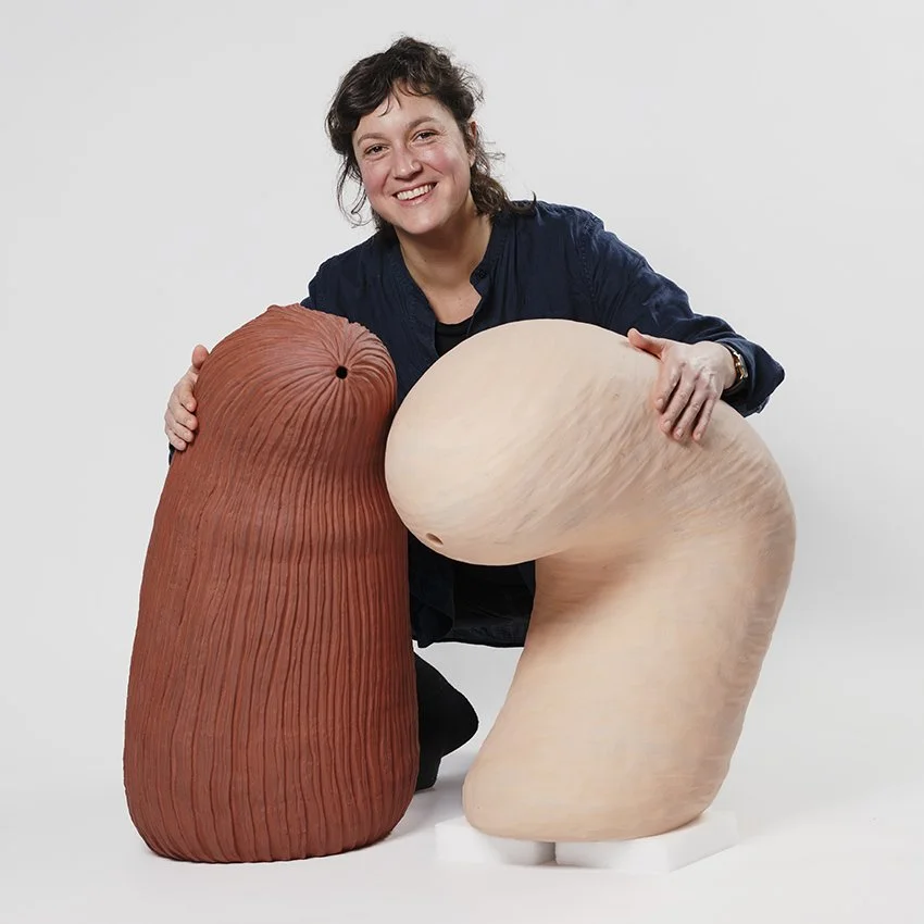 En kvinna ler och håller i två stora abstrakta skulpturer, en röda och en ljusbeige, mot en vit bakgrund.