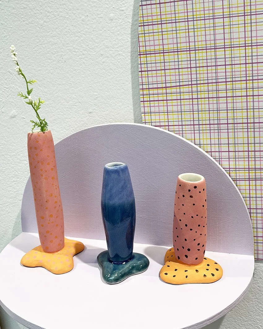 Tre konstnärliga keramiska vaser i olika färger och former, placerade på en vit yta med en bakgrund av en vit vägg och en grafiskt mönstrad tyg.
