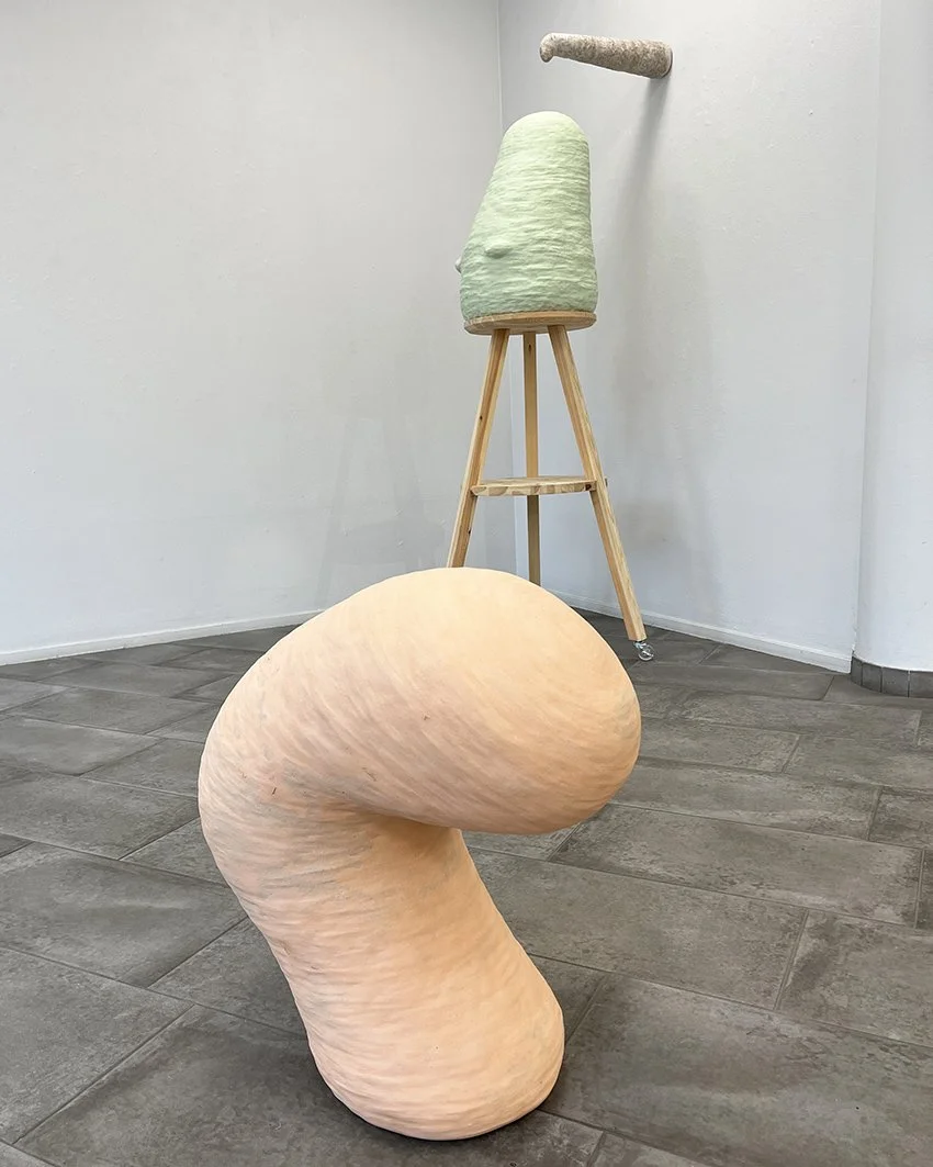 Skulptur av ett abstrakt, rosa, organliknande objekt i förgrund och ett trädstöd med ett grönt, keramiskt element på, i ett vitt rum med grå golv.