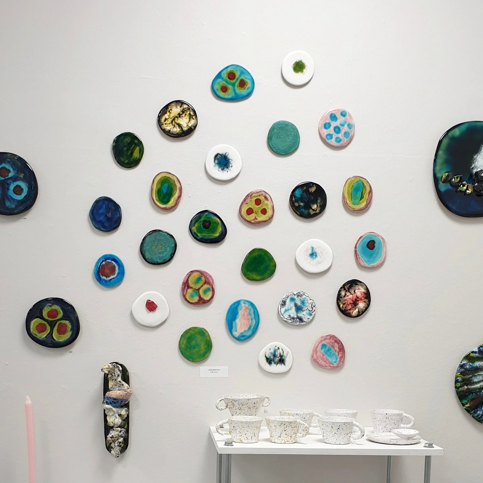 Galleri med färgglada, cirkelformade konstverk på vit vägg och vit bord med keramiska koppar och fat.