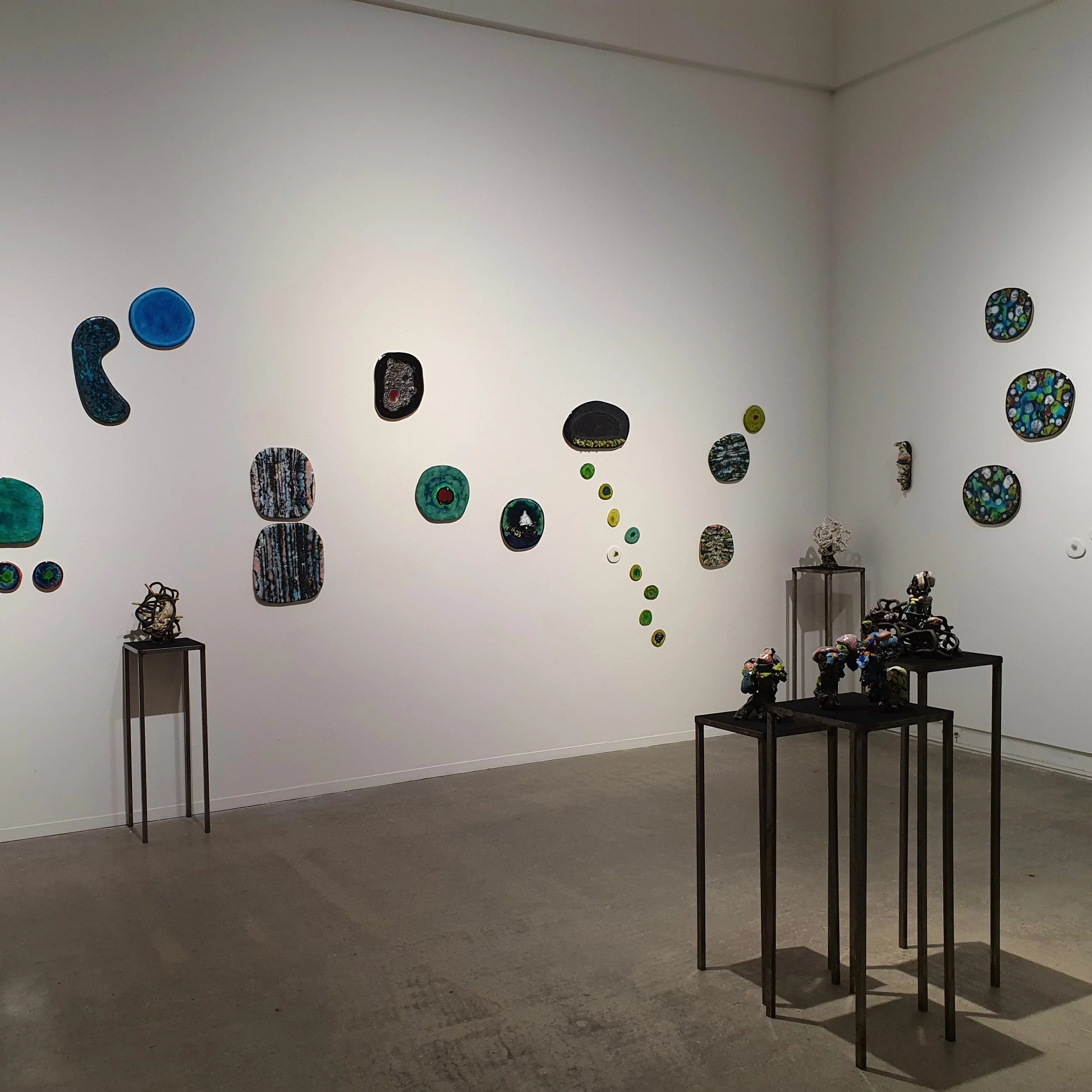 En konstutställning i ett galleri med väggar dekorera med många färgglada, abstrakta konstverk och skulpturer placerade på höga konsoler i mitten av rummet.