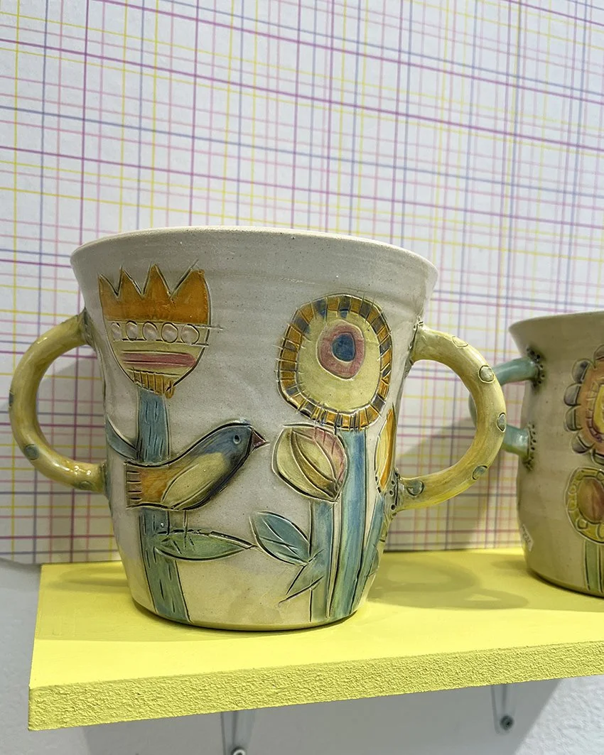 Ceramicmugg med blomster- och fågelmotiv på en gul hylla, mot en bakgrund av rutigt mönster.