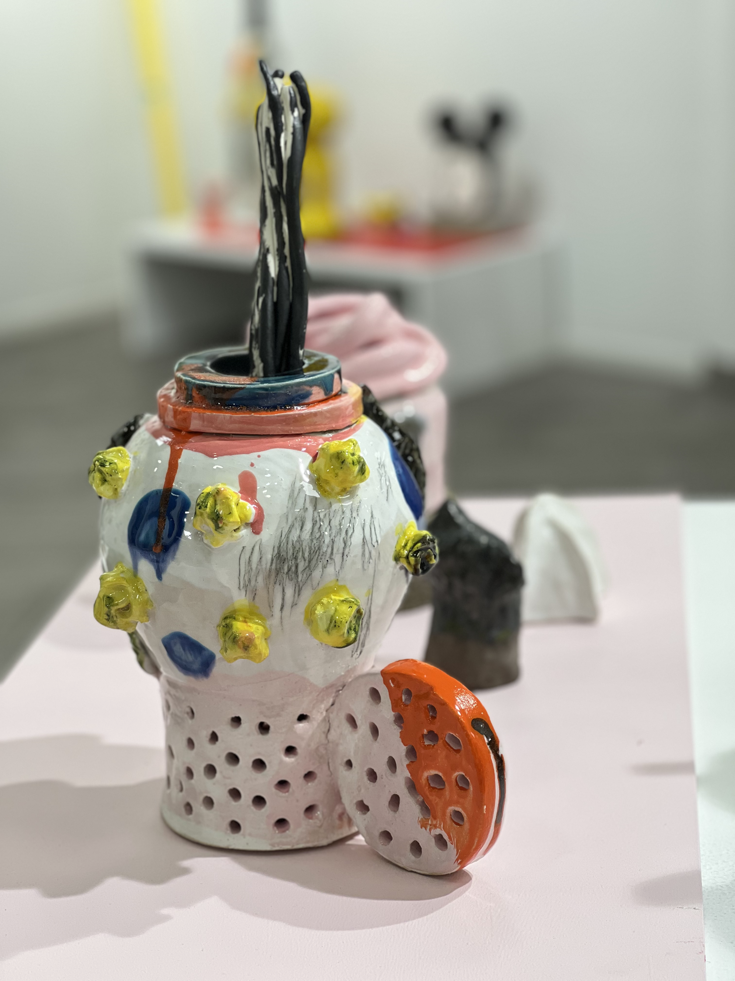 Färgglad konstnärlig skulptur av keramikkopp med dekorativa element som popcorn, ett blomsterblad och svarta strån, placerad på en ljus yta i ett galleri eller utställningsrum.