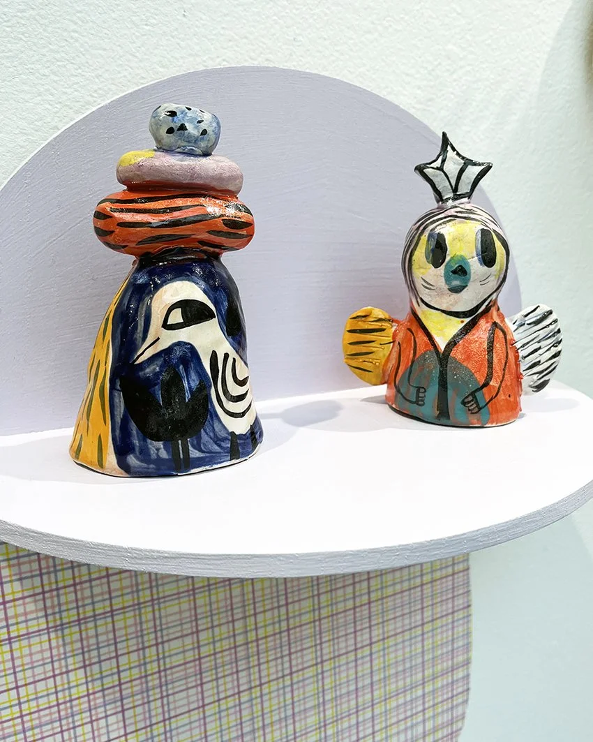 Färggranna, handgjorda keramikfigurer av djur, här en med en stack av fyra stenar ovanpå samt en med ett julhatt, placerade på en vit väggmonterad hylla.