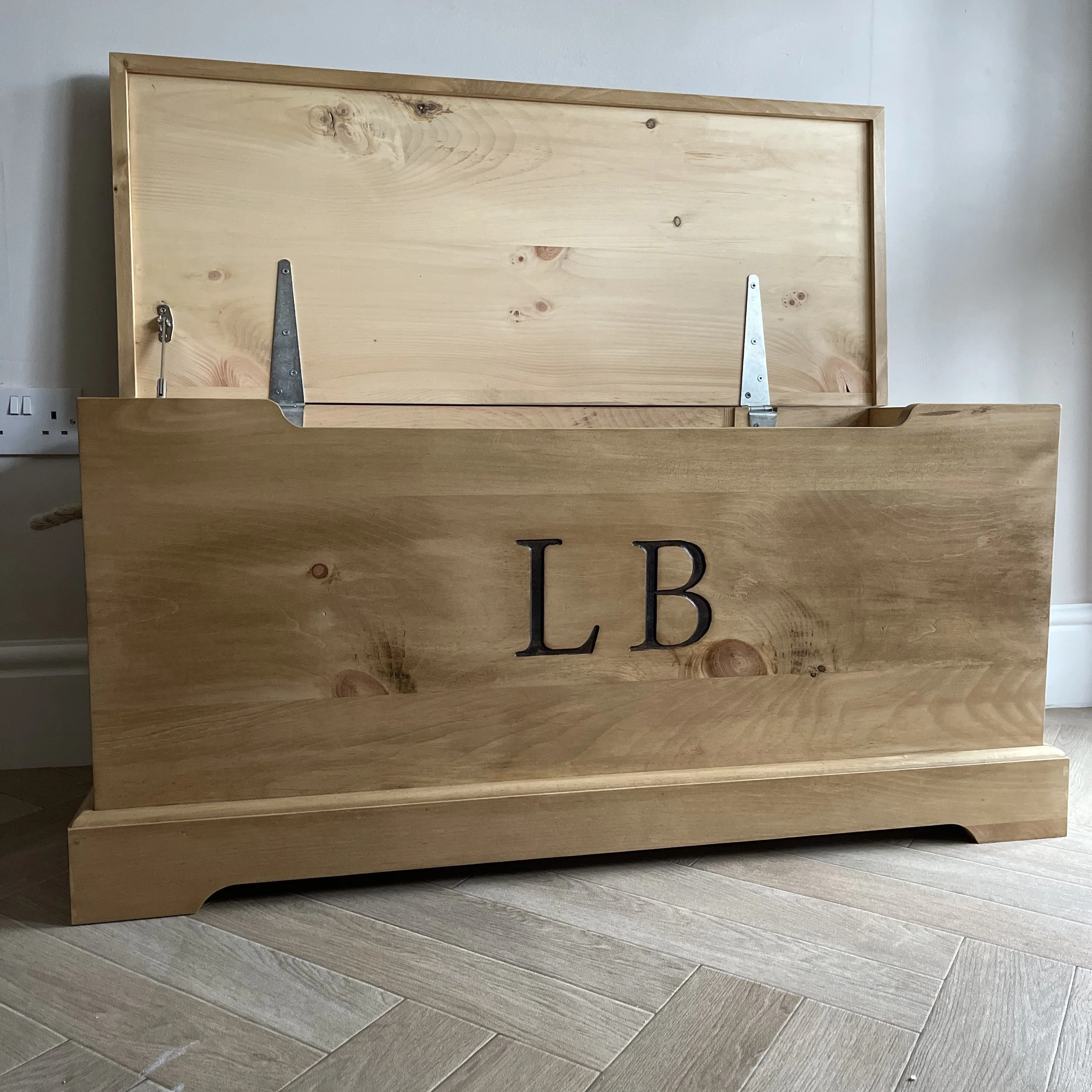 Handmade wooden toy box with open lid and personalised engraving, placed on herringbone wooden floor.