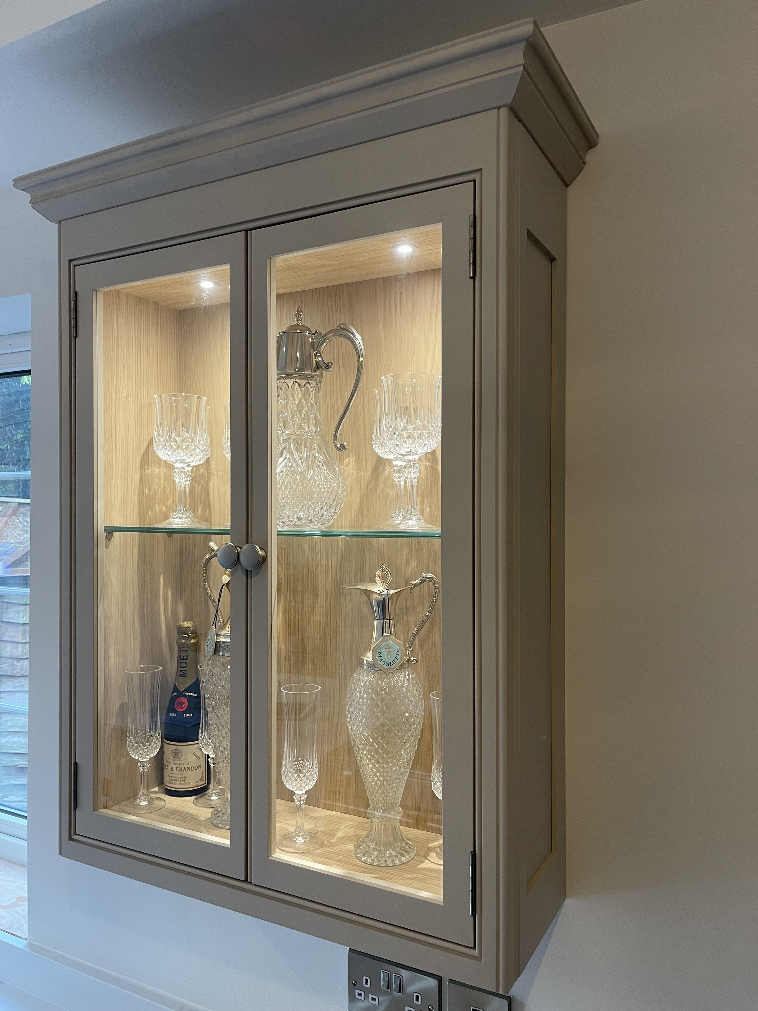Bespoke kitchen display cabinet with glass doors showcasing crystal glassware and a champagne bottle.