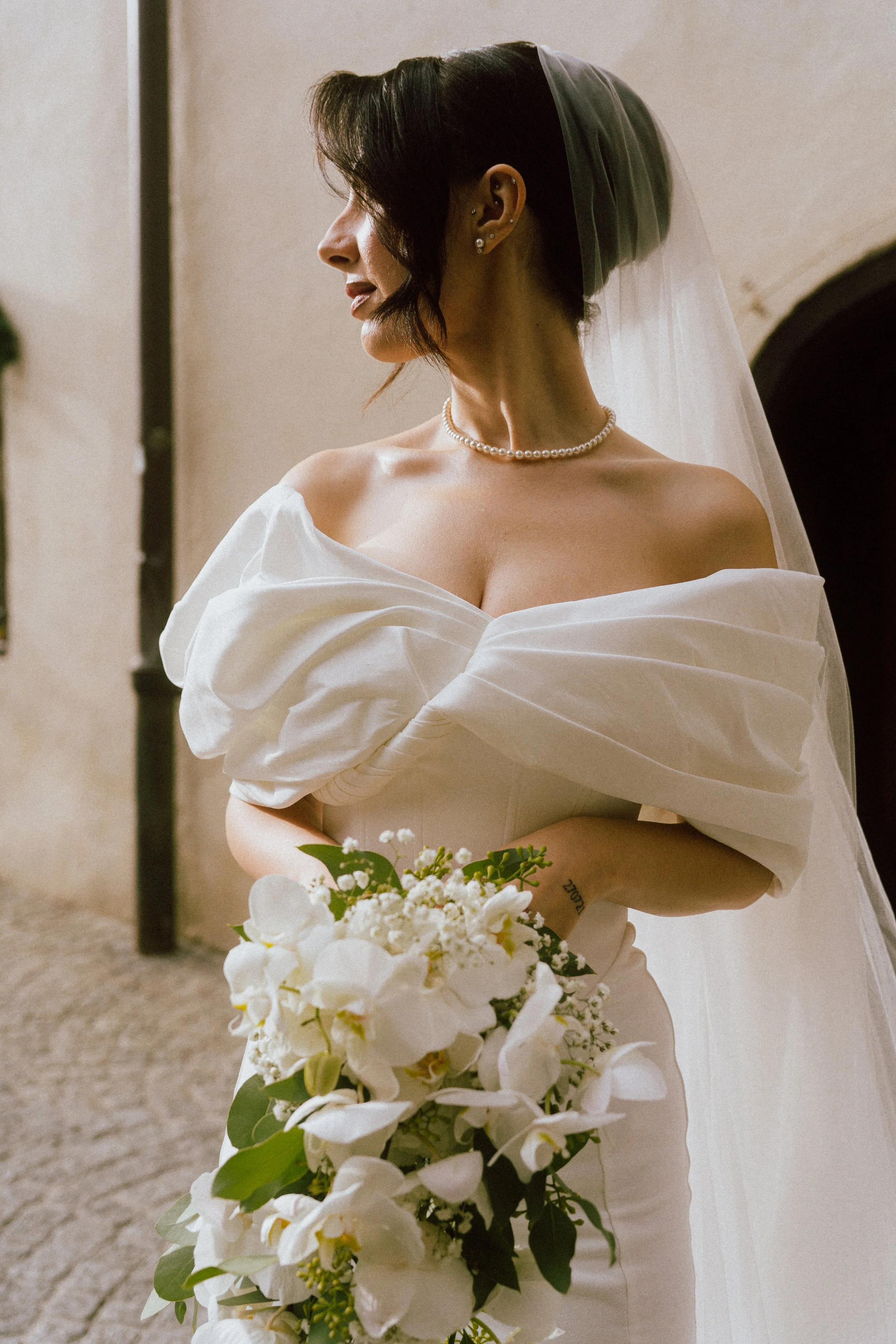 Frau in Hochzeitskleid mit Schleier und Perlenkette hält einen Blumenstrauß aus weißen Orchideen und kleinen weißen Blumen.