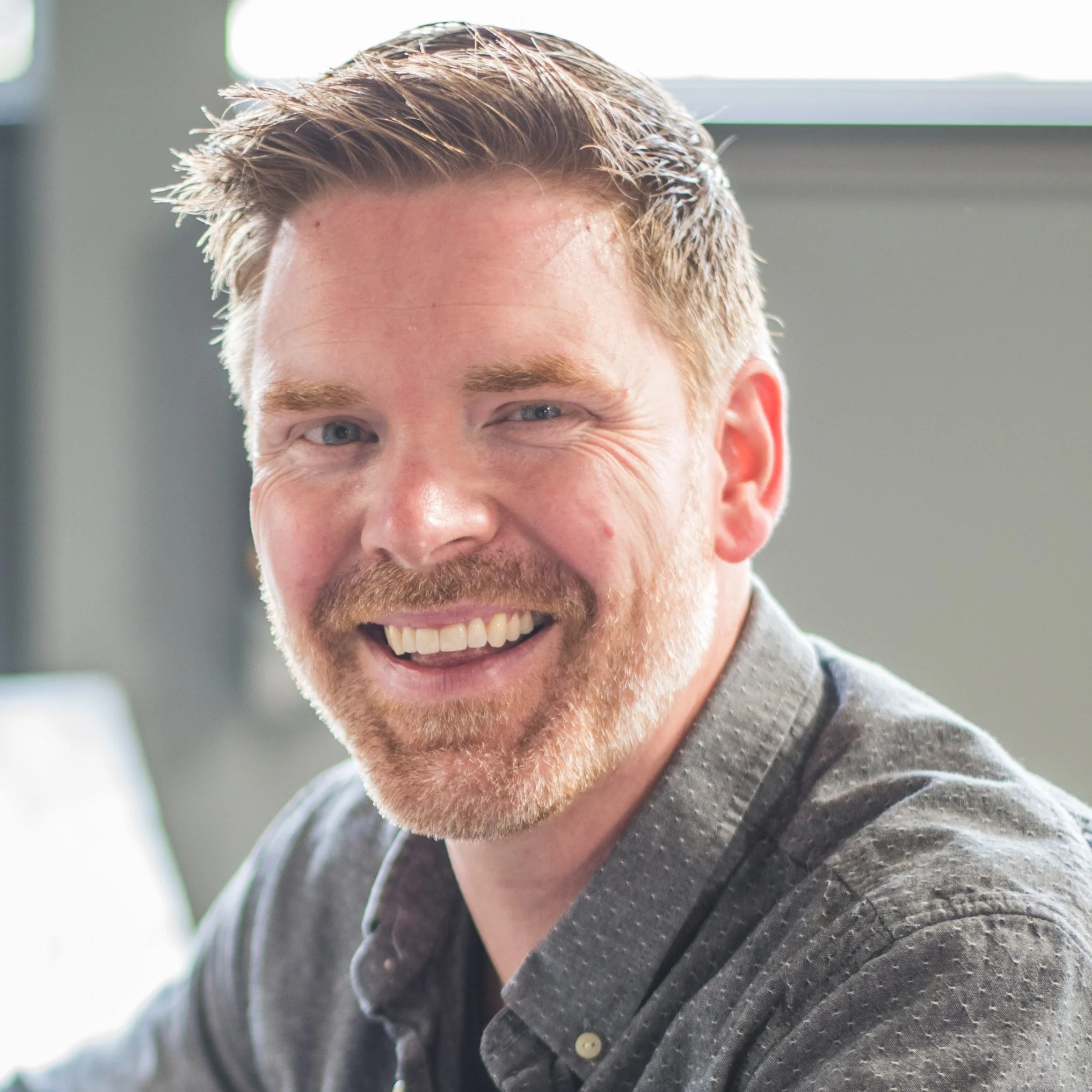 A smiling man with ginger hair and a beard, wearing a gray button-up shirt, in an office setting.
