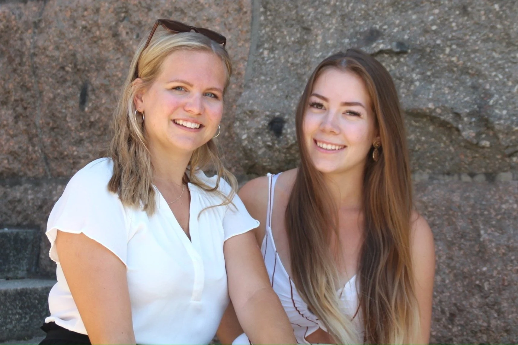 Zwei lachende junge Frauen sitzen nebeneinander vor einem Steinmauer, machen ein Foto und genießen den Tag.
