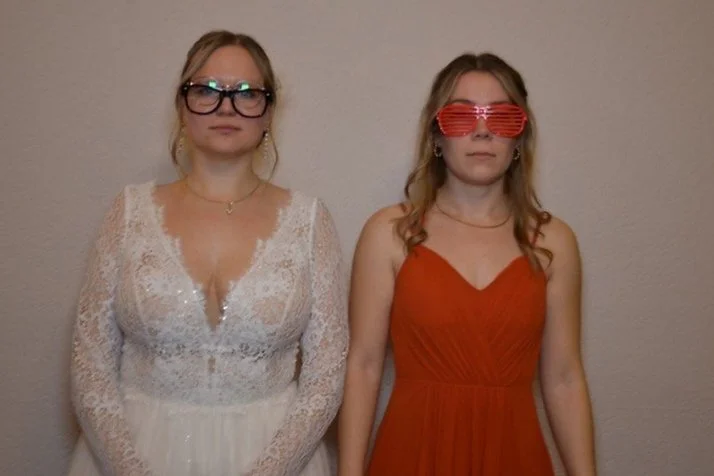 Zwei junge Frauen stehen vor einer weißen Wand, tragen Partyoutfits und bunte Sonnenbrillen, eine in Weiß und die andere in Rot.