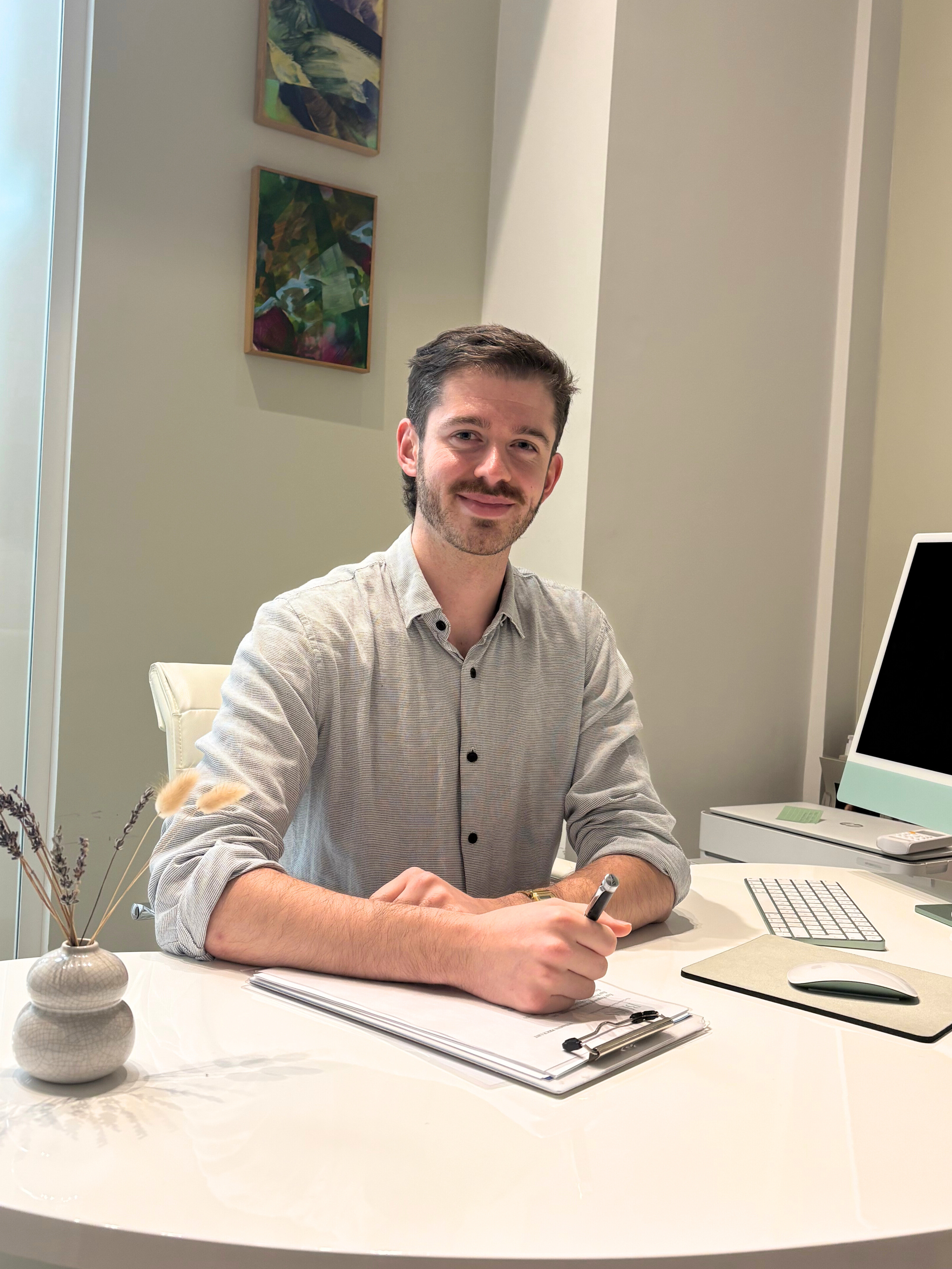 A man sitting at a desk in an office, smiling, with a pen in his right hand, a clipboard, a computer, and a vase with dried flowers on the desk.