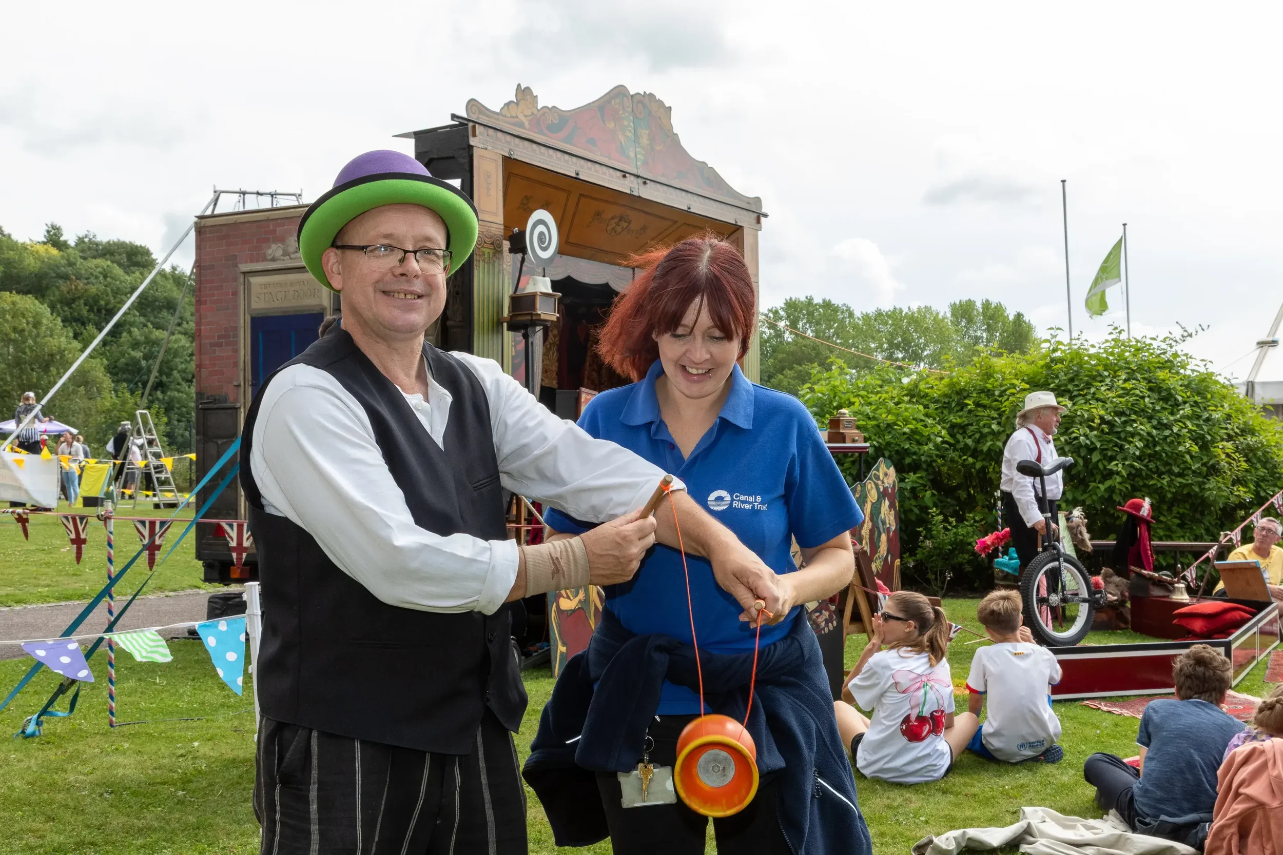 A circus performer dresses in black trousers, white shirts, black waistcoat and green and purple bowler hat helping a young woman practice diabolo, she is wearing a blue polo shirt. They are both outdoors at an event.