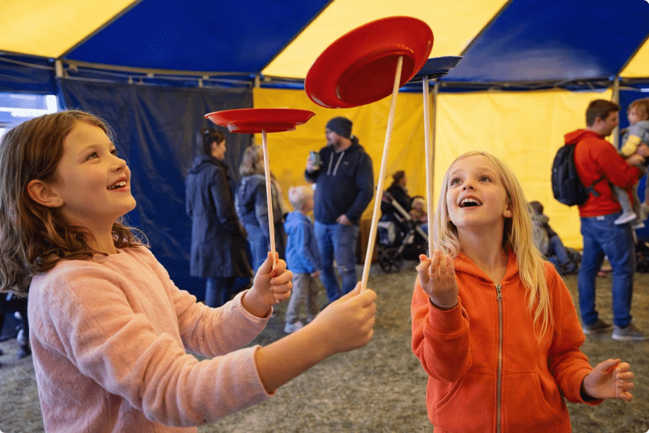 Two girls spinning red plates on sticks inside a blue and yellow circus tent, smiling as people watch in the background.