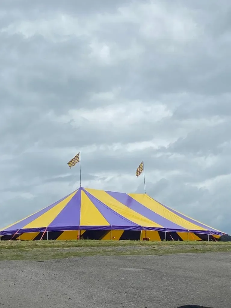 A large striped circus tent with blue and yellow panels, set up outdoors on a grassy field under a cloudy sky, with flags on top.