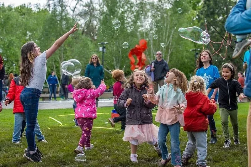 a group of 10 young children laughing and playing in a park with floating bubbles