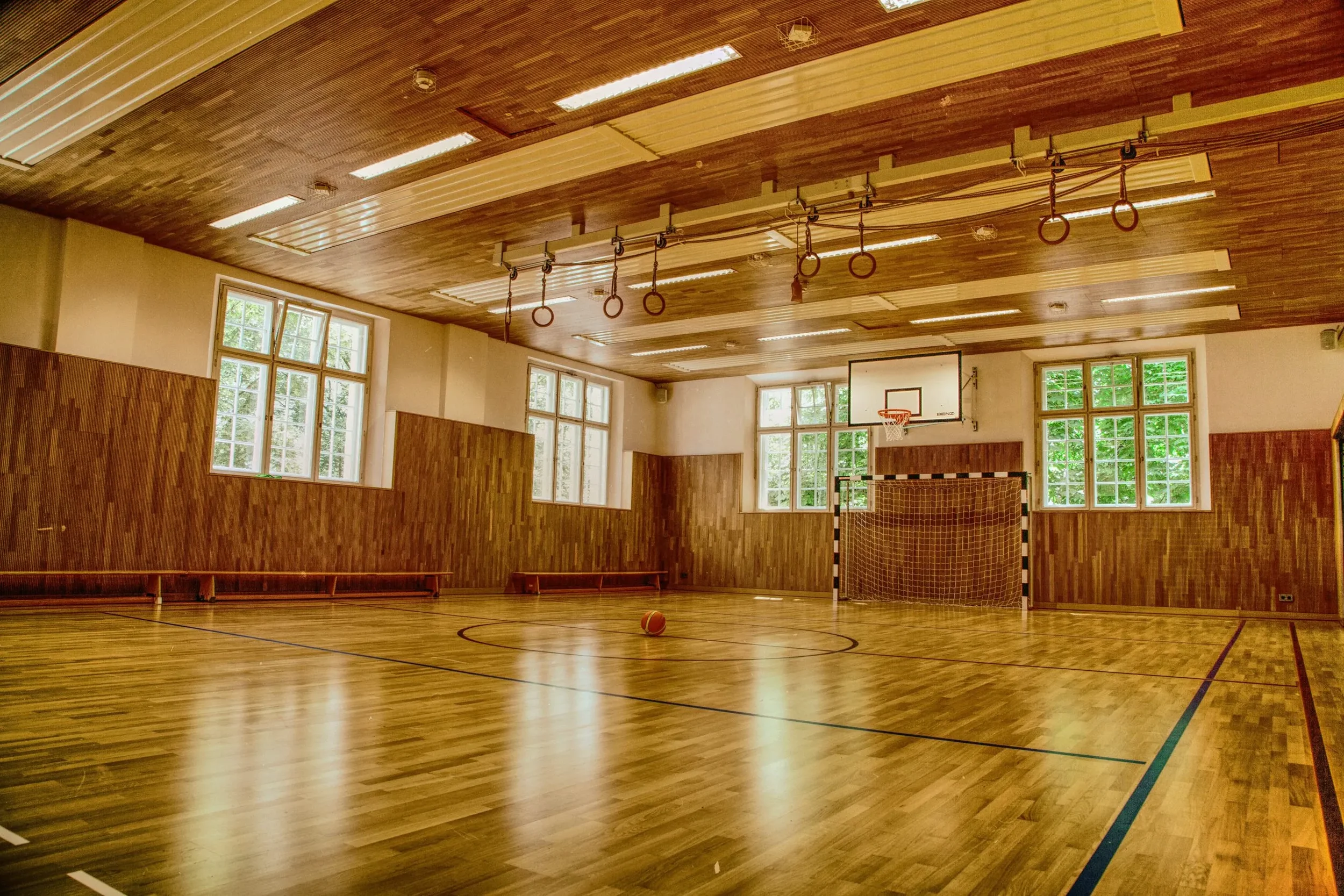 Empty indoor basketball court with wooden floors, basketball hoop, and gymnastic rings hanging from the ceiling, illuminated by natural light through large windows.