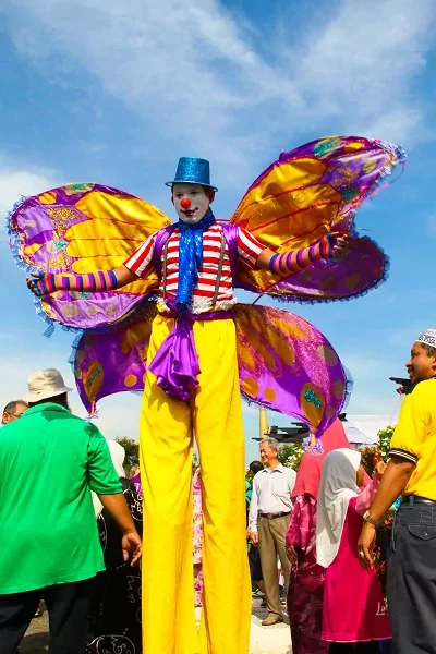 brightly coloured stilt walker dressed like a butterfly with yellow trousers, a red nose and a blue top hat