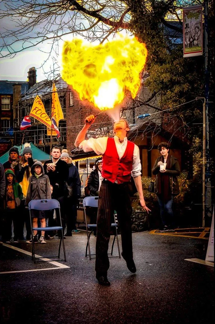 Circus fire performer in red waistcoat and white shirt breathing fire in front of a crowd at a UK event