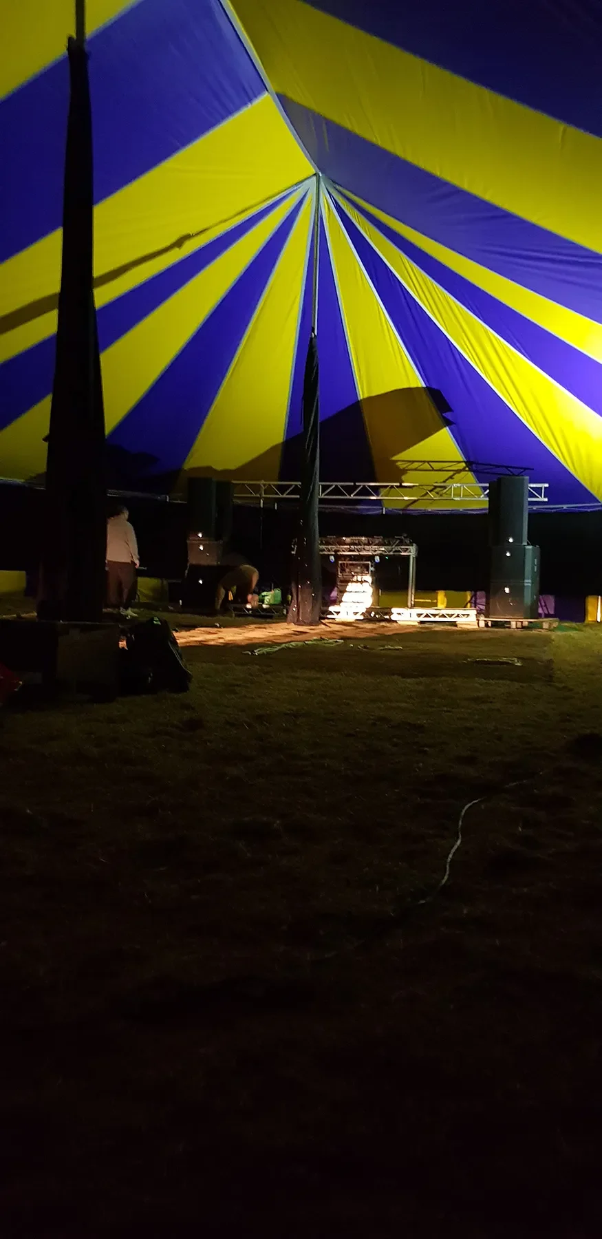 Nighttime scene of a partially assembled large yellow and purple striped tent with a stage setup underneath, and a few people working on the setup.