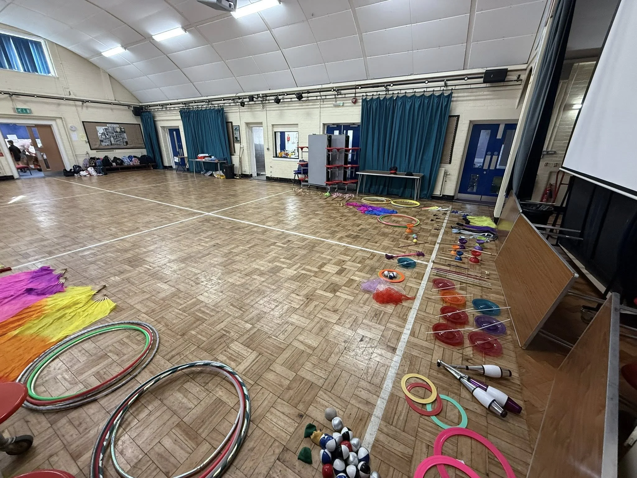 Large indoor sports hall with wooden floor and colourful circus equipment—hula hoops, juggling clubs, plates, rings, and scarves—laid out along the edge ready for a workshop