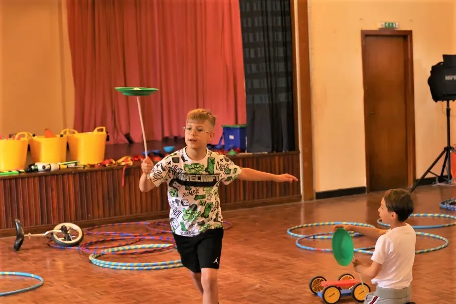 2 young boys in a school gymnasium with circus equipment all over the floor playing with spinning plates