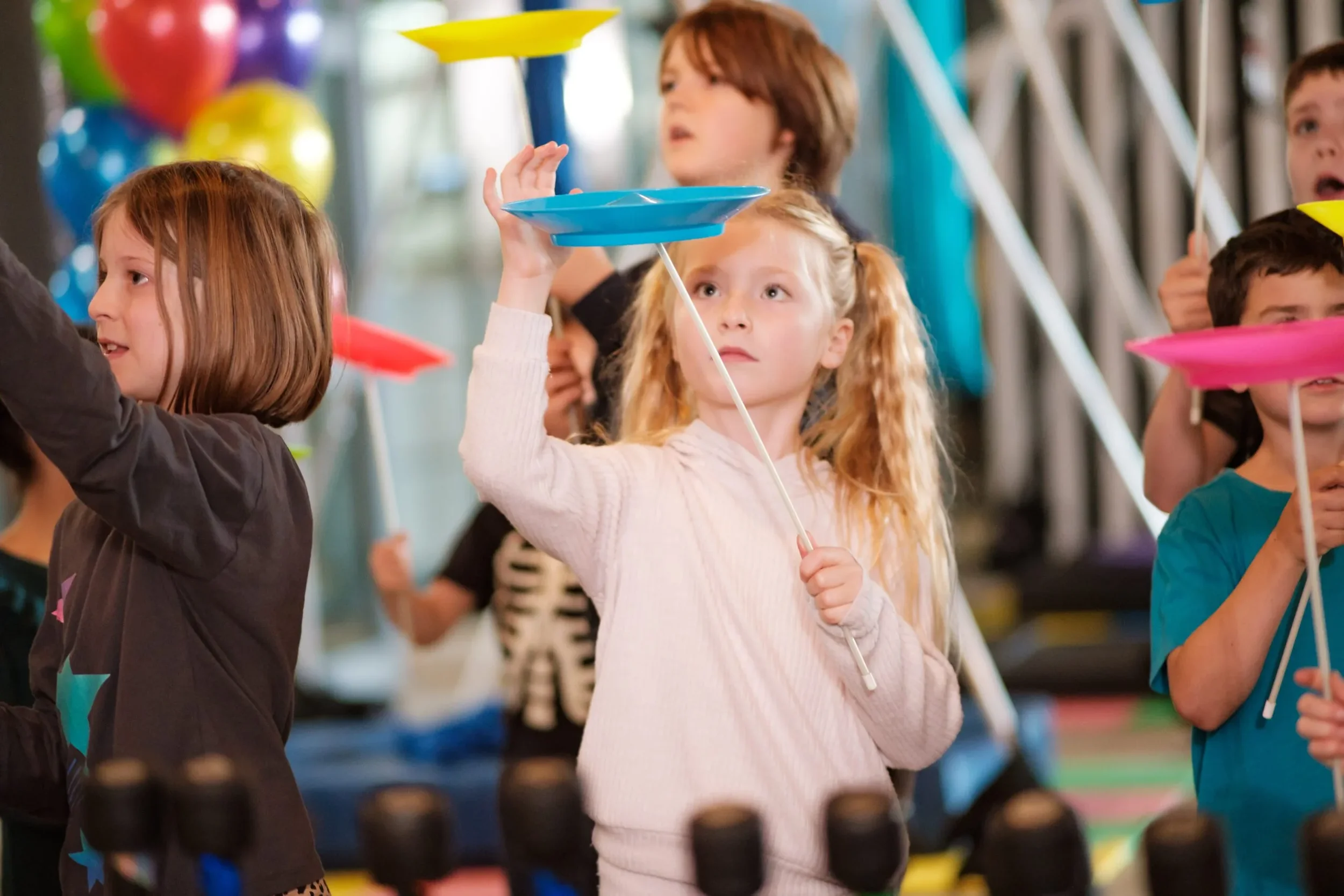 a small group of young children playing with spinning plates focusing on a young girl at the front with a light pink jumper playing with a blue spinning plate