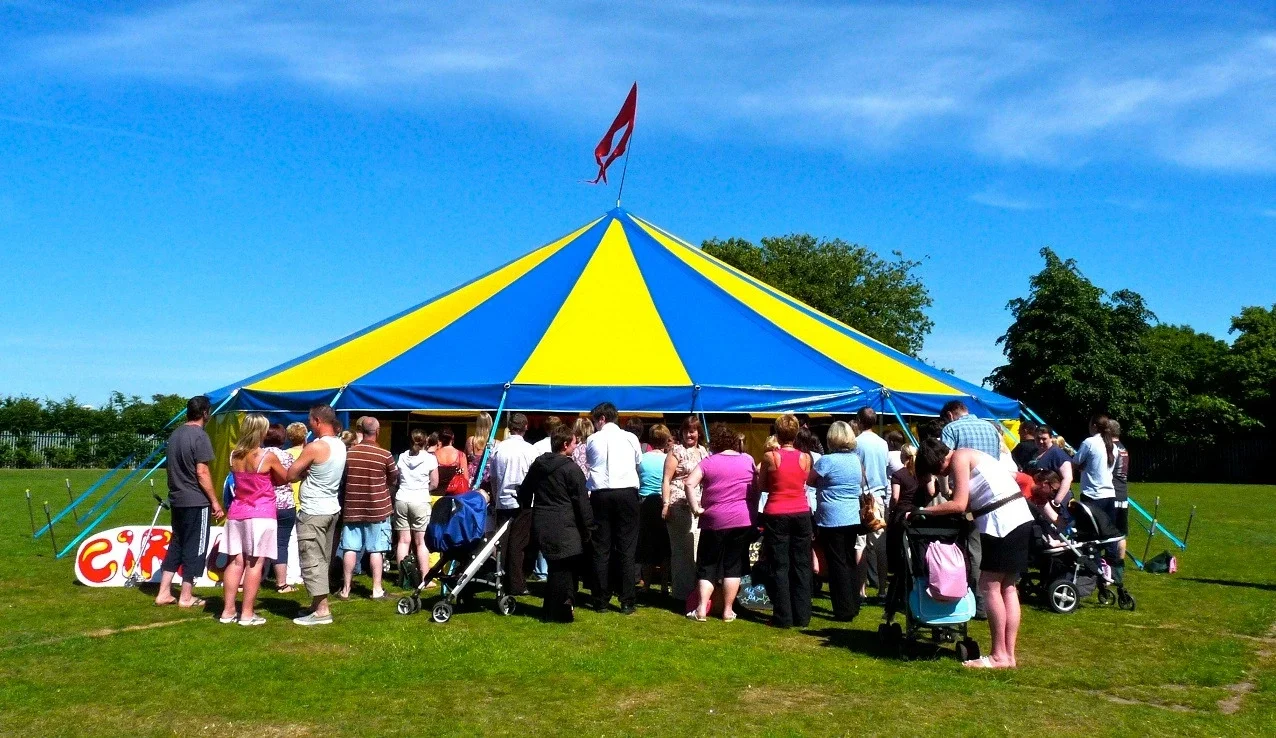 single pole blue and yellow circus tent with a large crowd of people around set on grass with bright blue sky