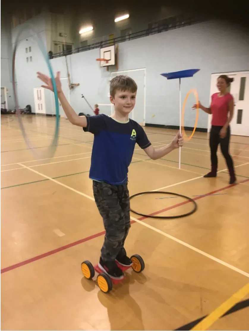 young boy dressed in blue t shirt and camouflage trousers playing with a hola hoop, fun wheels and spinning a plate in a gymnasium with a adult watching and encouraging from the right side