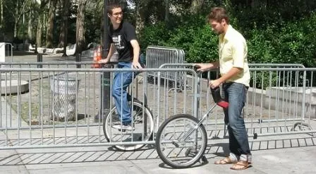 2 people with unicycles one riding one and one just learning with steel railings