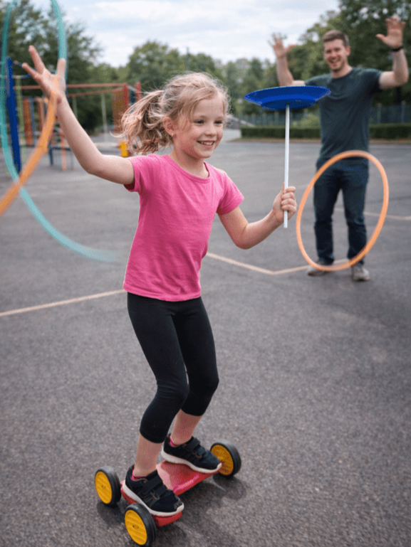 Girl in a pink top riding a red peddle-go on an outdoor playground, spinning a blue plate while one adult cheers in the background.