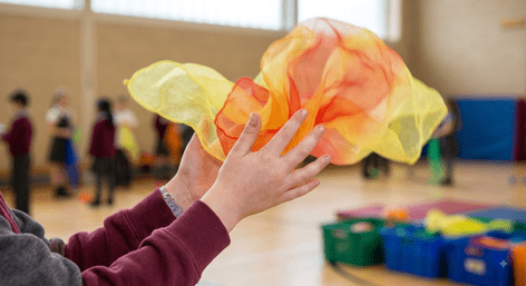 A close-up view of a child's hands catching a yellow and orange juggling scarf in a gymnasium