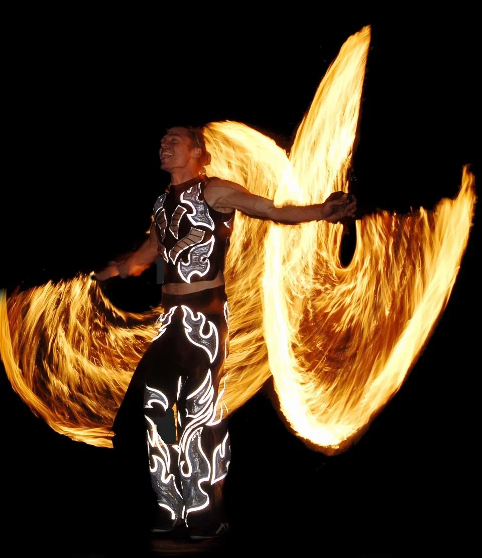 Professional circus fire performer spinning large fire fans creating dramatic arcs of flame against a dark night sky at a UK event