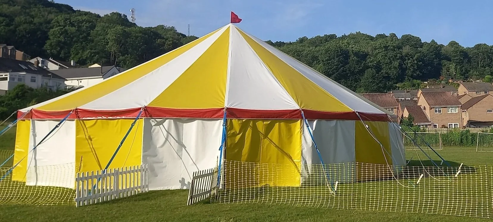 A yellow and white striped circus tent set up on a grassy field with a small white picket fence and a yellow plastic barrier around it, against a backdrop of residential houses and green trees.