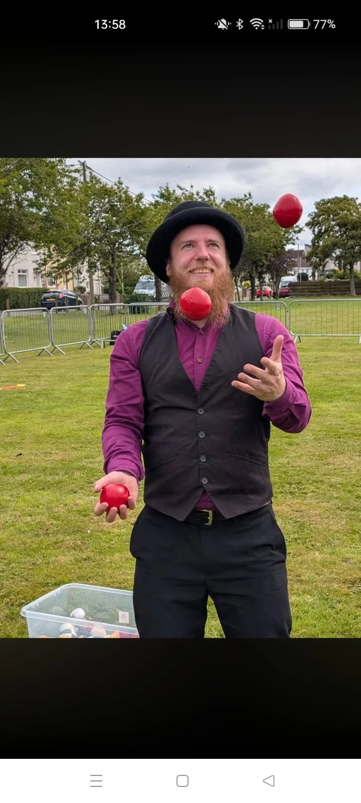 a juggler in a black bowler hat purple shirt and waistcoat standing in a field juggling 3 red balls