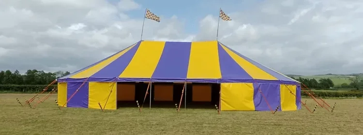 A large yellow and blue striped circus tent set up on a grassy field with cloudy sky overhead.