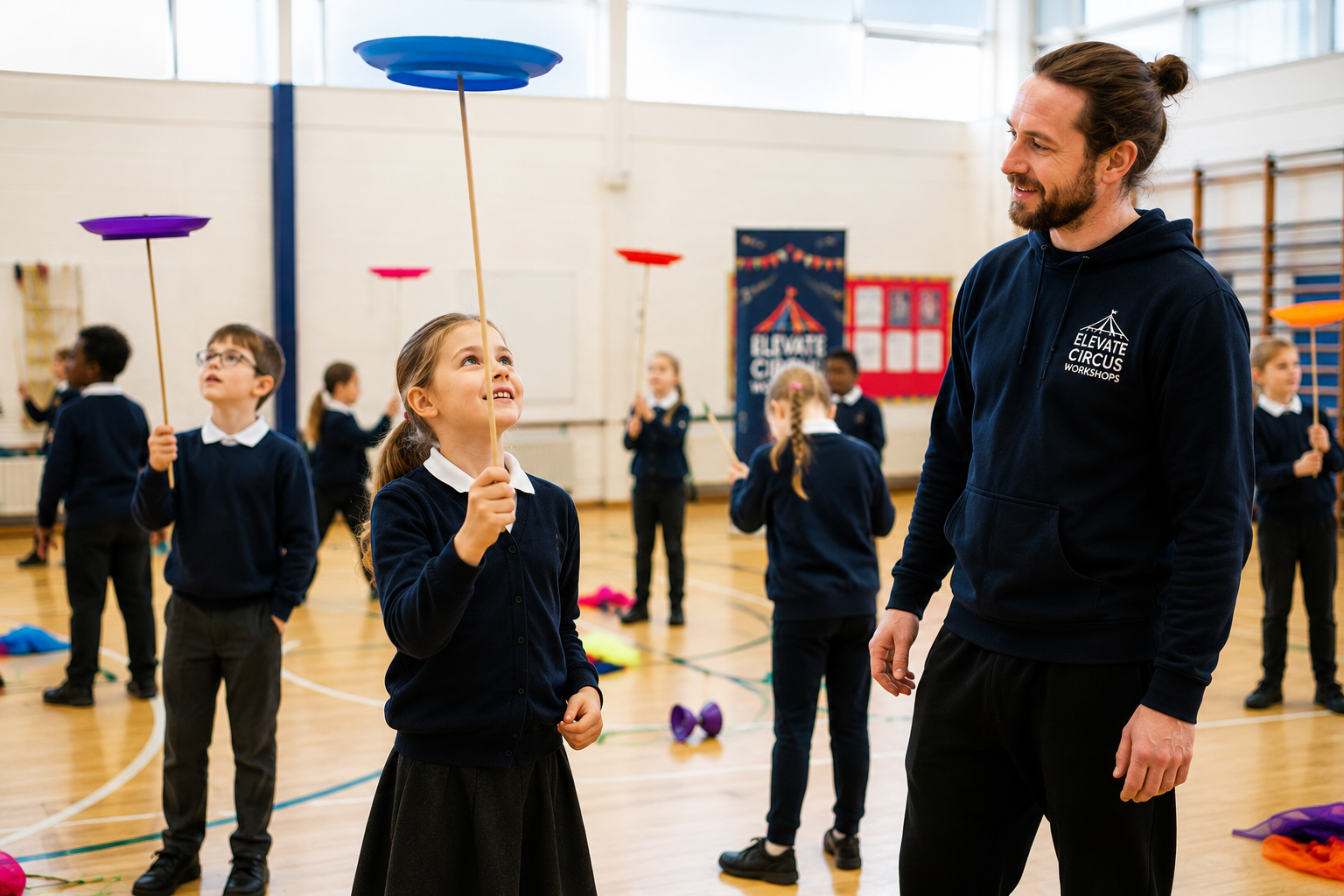 Primary school child in school uniform smiling while concentrating on spinning a plate on a stick during a circus skills workshop, with a professional tutor offering encouragement in a bright school sports hall.