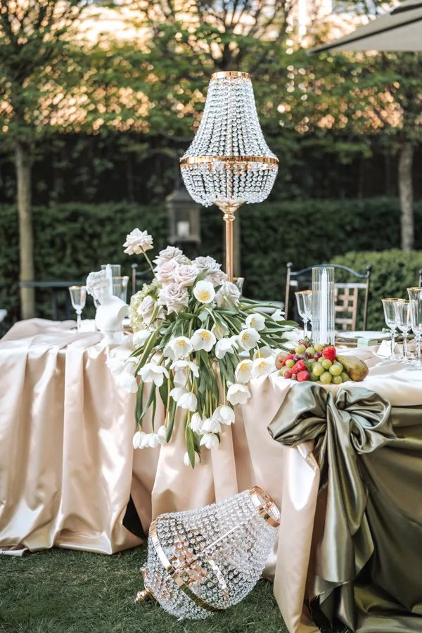 Table de mariage élégante avec centre de fleurs blanches, chandelier en cristal et fruits, située en plein air avec un fond de feuillage et de ciel au coucher du soleil