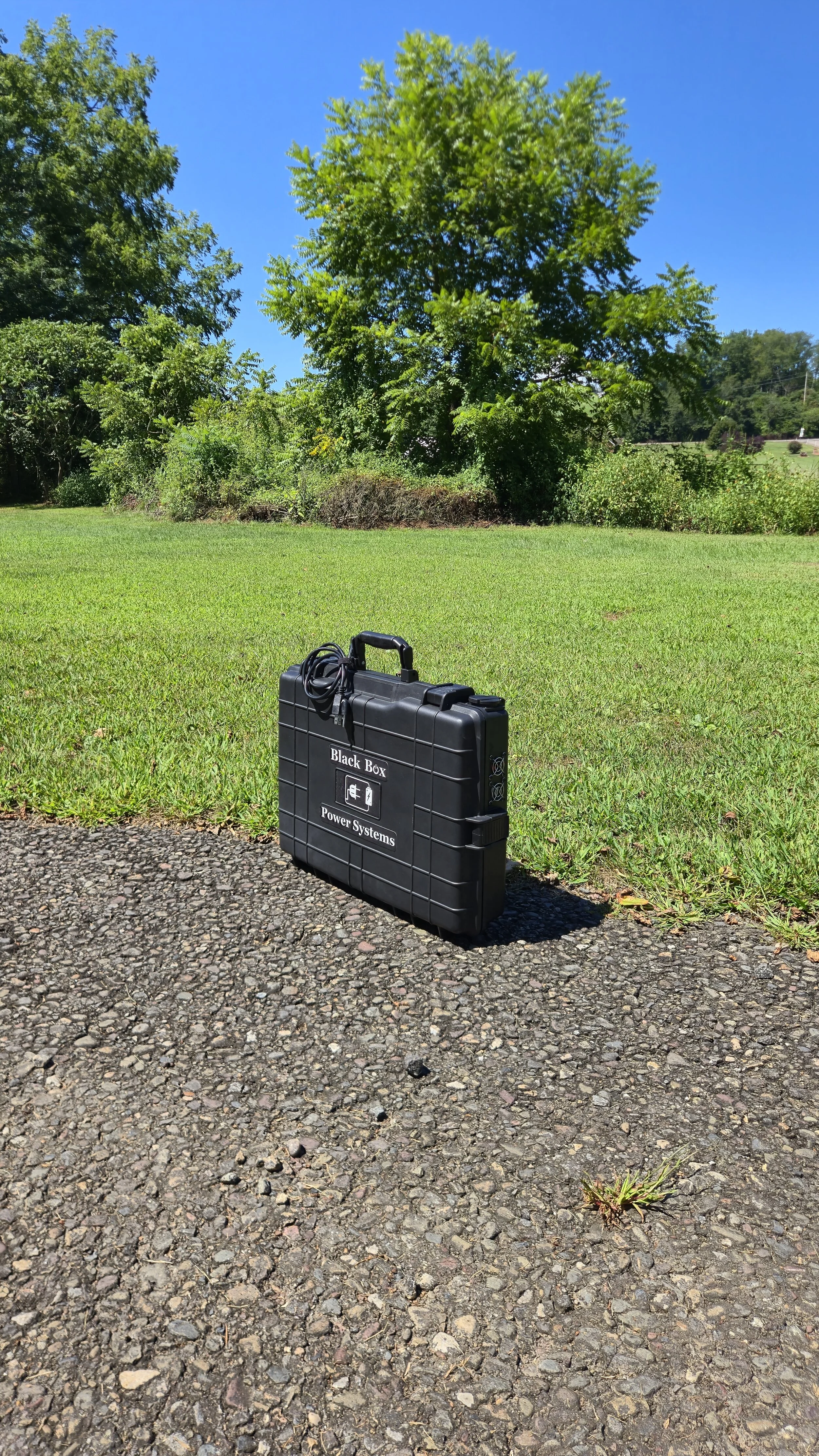 Black portable power system case labeled 'Black Box Power Systems' on a gravel surface outdoors, with green grass, trees, and a clear blue sky in the background.