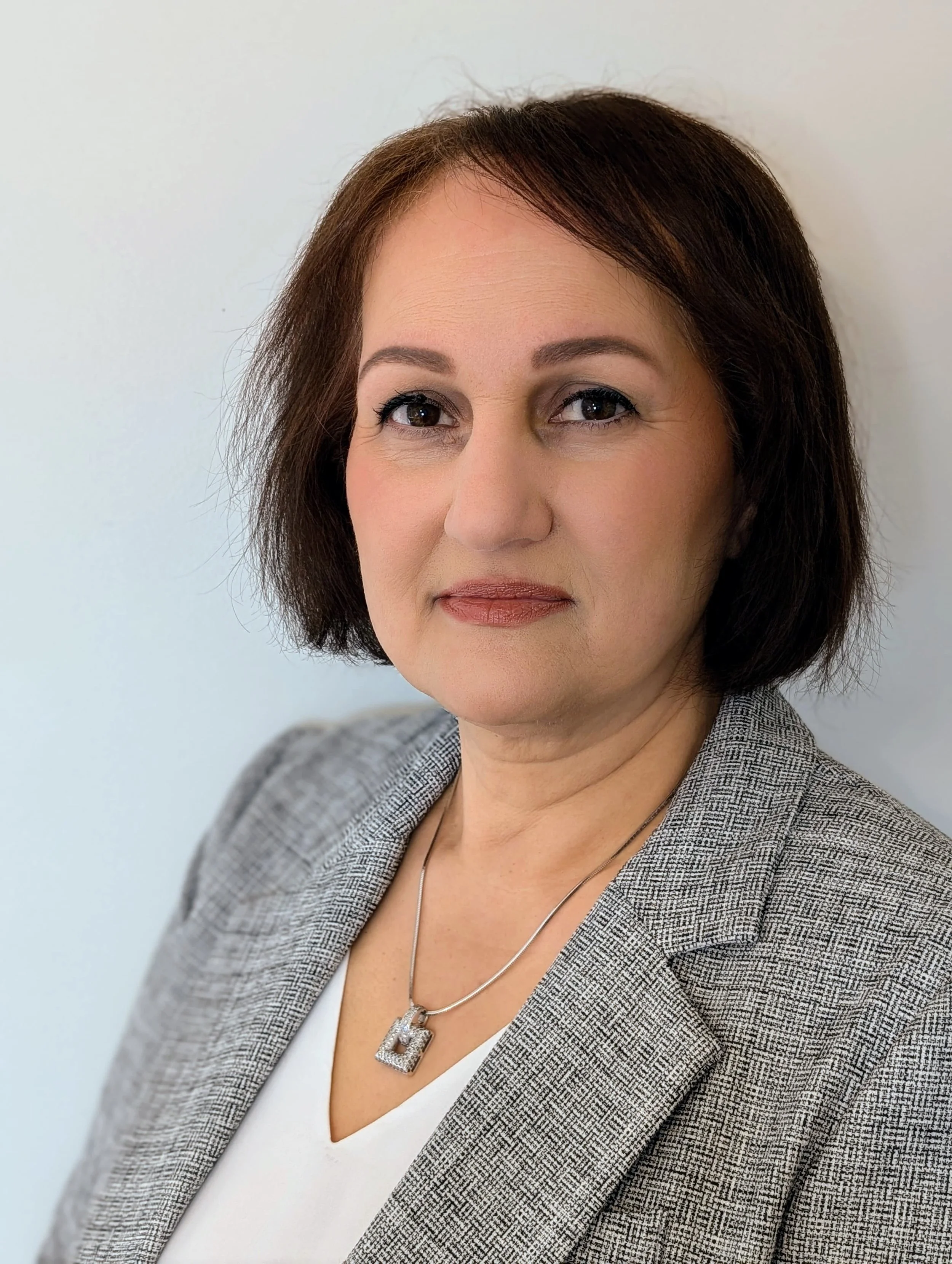 A woman with short brown hair and light skin, wearing a gray blazer over a white top, and a silver necklace with a decorative pendant, against a plain light background.