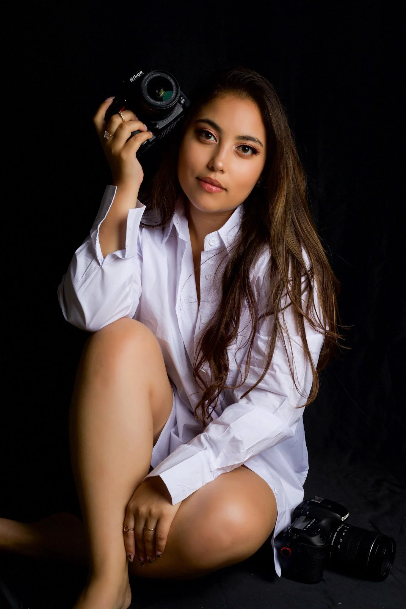 A young woman with long brown hair, wearing a white shirt, sitting against a black background, holding a Nikon camera on her shoulder, with another camera on the ground beside her.