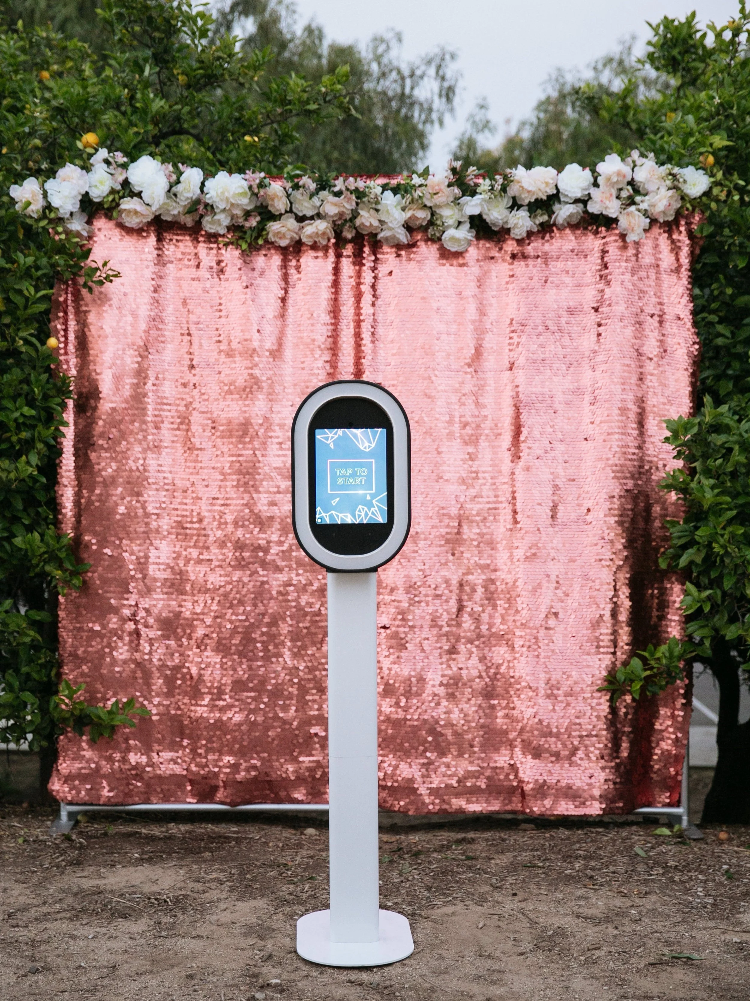 A photo of an outdoor event setup featuring a pink, sequined backdrop with a floral arrangement of white and blush roses along the top. In front of the backdrop is a digital kiosk with a touch screen display that reads 'TAP TO START'.