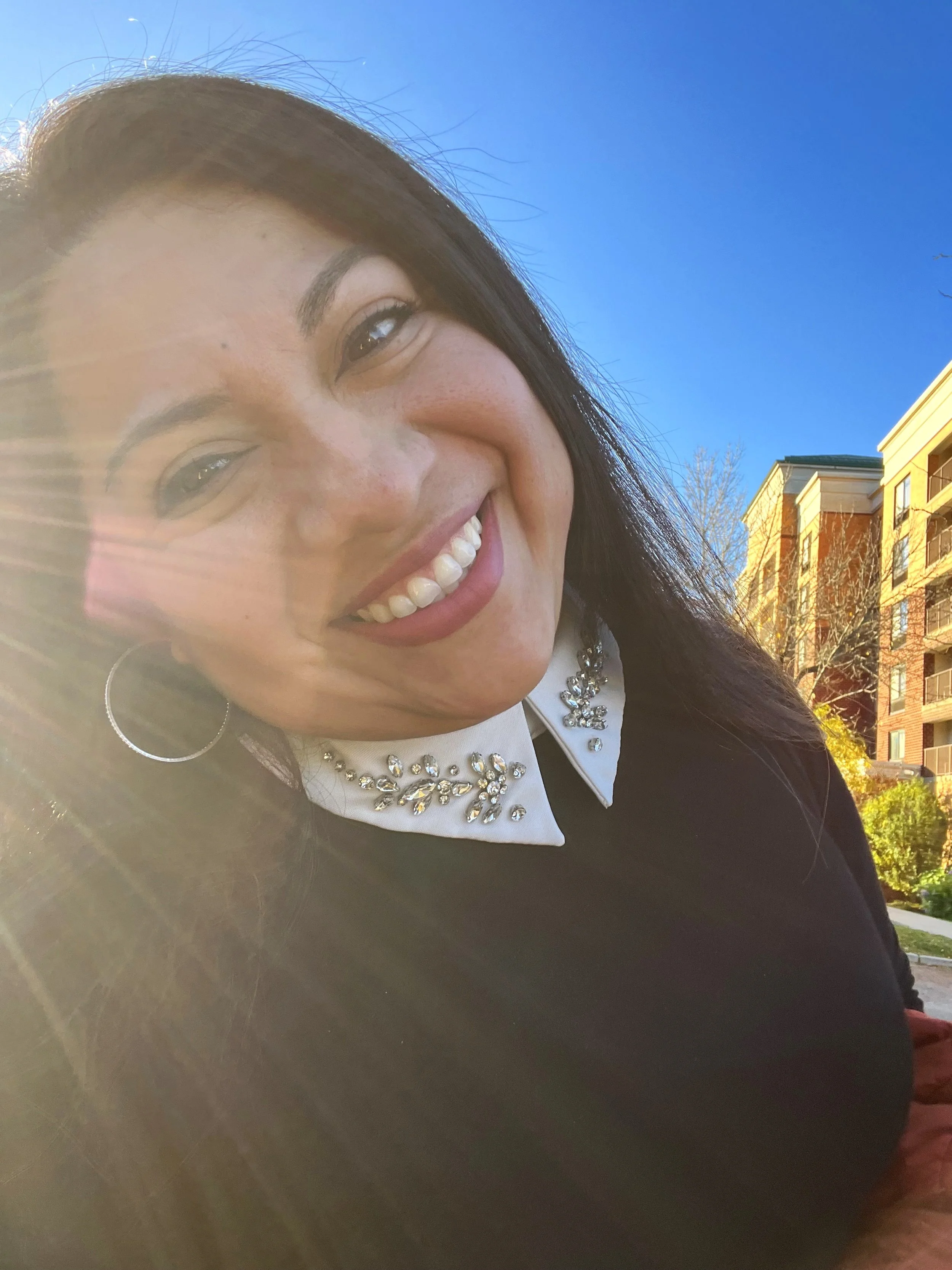 Close-up of a smiling woman with dark hair, wearing a black top and a white collar with rhinestone details, outdoors on a sunny day, with buildings and blue sky in the background.