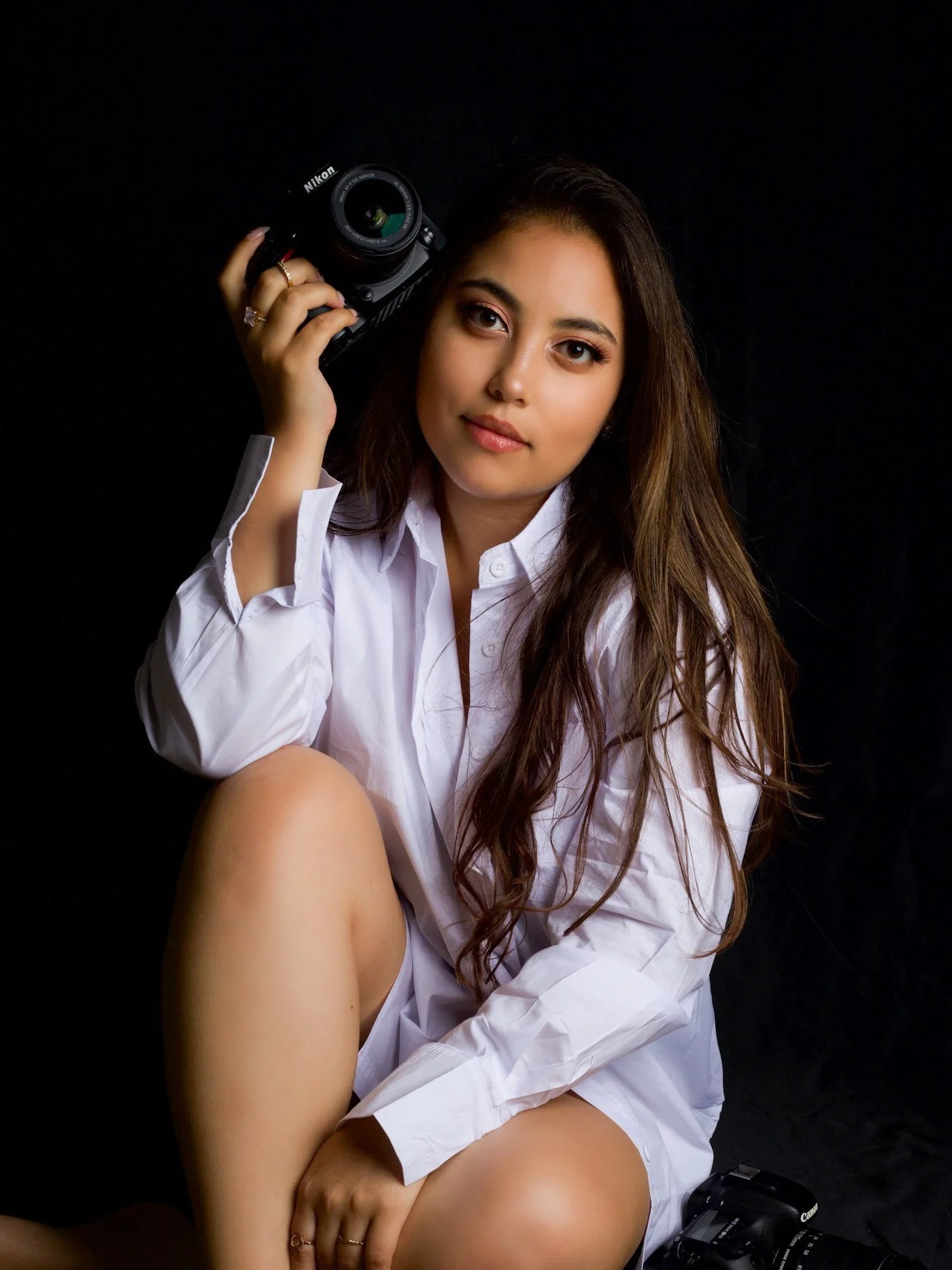 A young woman with long brown hair sitting against a black background, wearing a white shirt. She holds a Nikon camera near her head with her right hand and rests her left hand on her leg. She has a neutral expression and is looking directly at the camera.