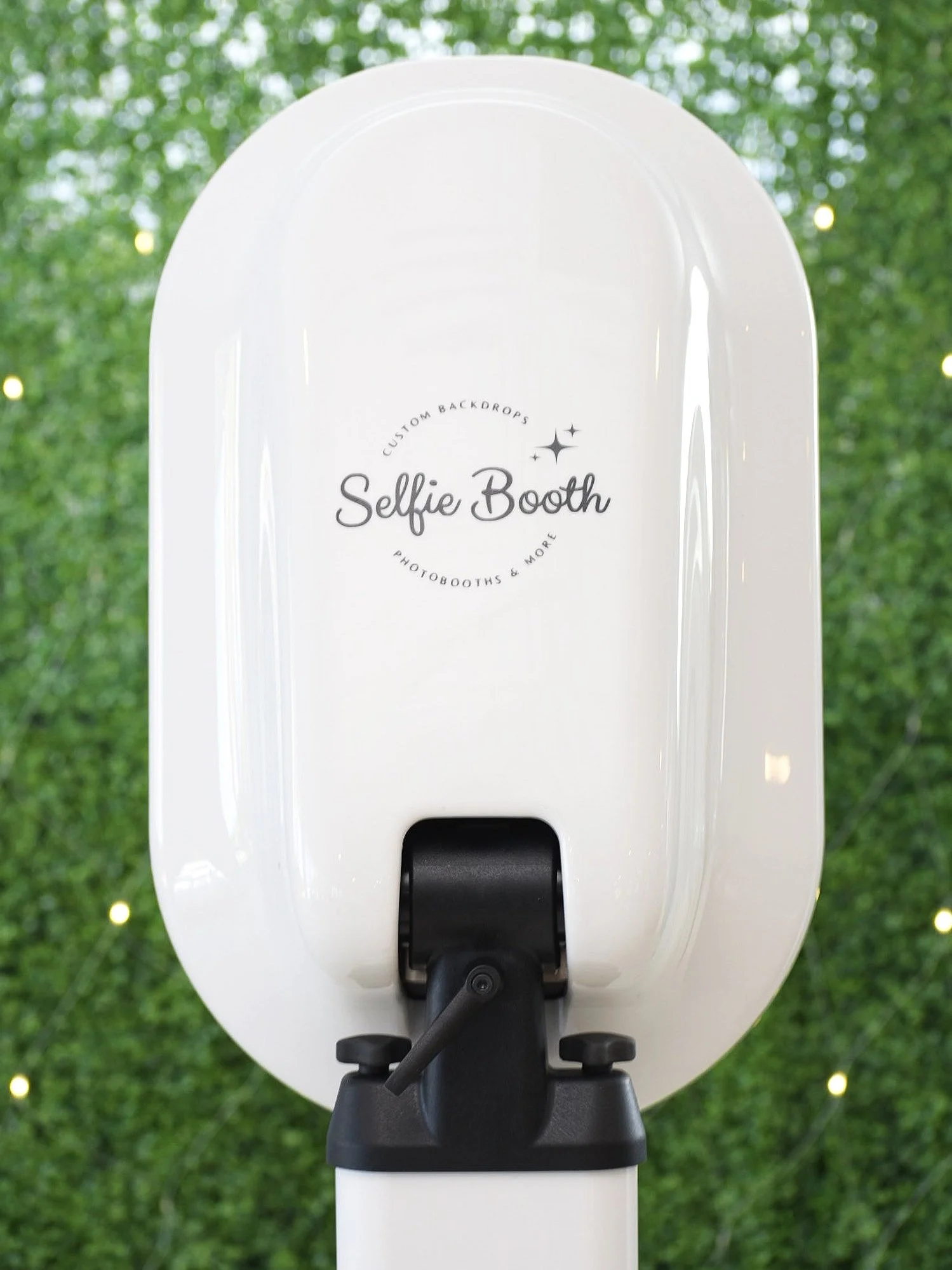 Close-up of a white selfie photo booth with black components, set outdoors with green aspen leaves and small string lights in the background