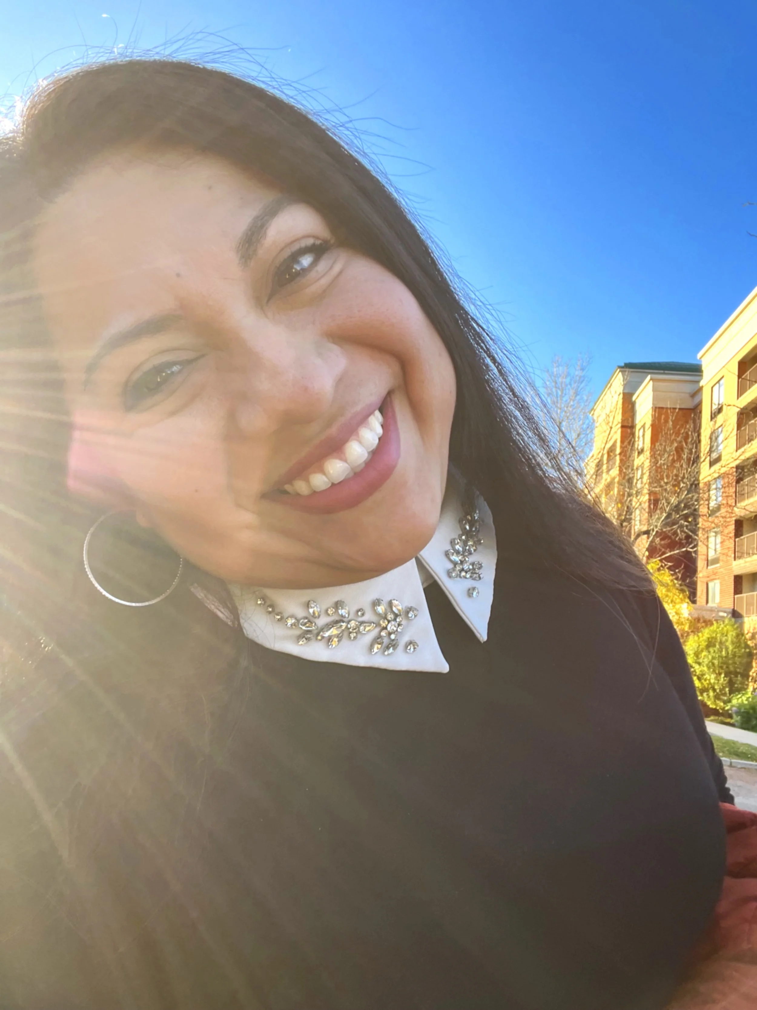 A woman smiling outdoors, wearing a white shirt with a bejeweled collar and large hoop earrings, with buildings and a clear blue sky in the background.