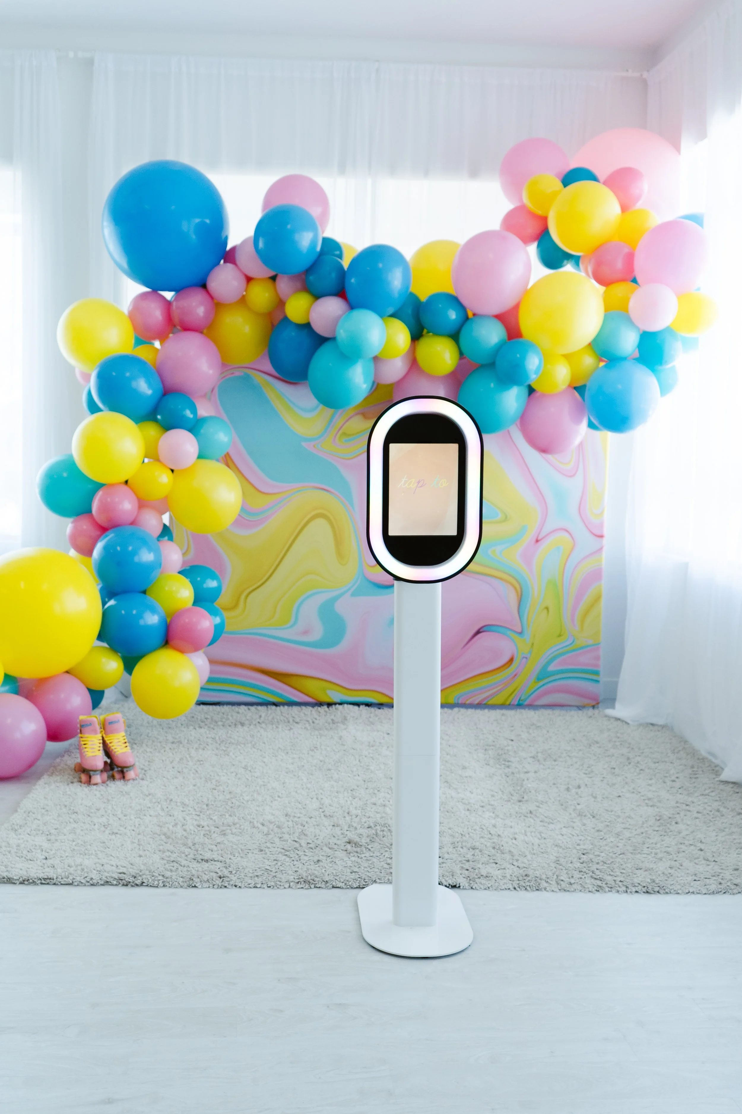 Colorful balloon decor with pink, blue, and yellow balloons arranged on a backdrop with pastel swirl pattern in a room with white curtains and beige carpet, featuring a touch screen kiosk in the foreground.