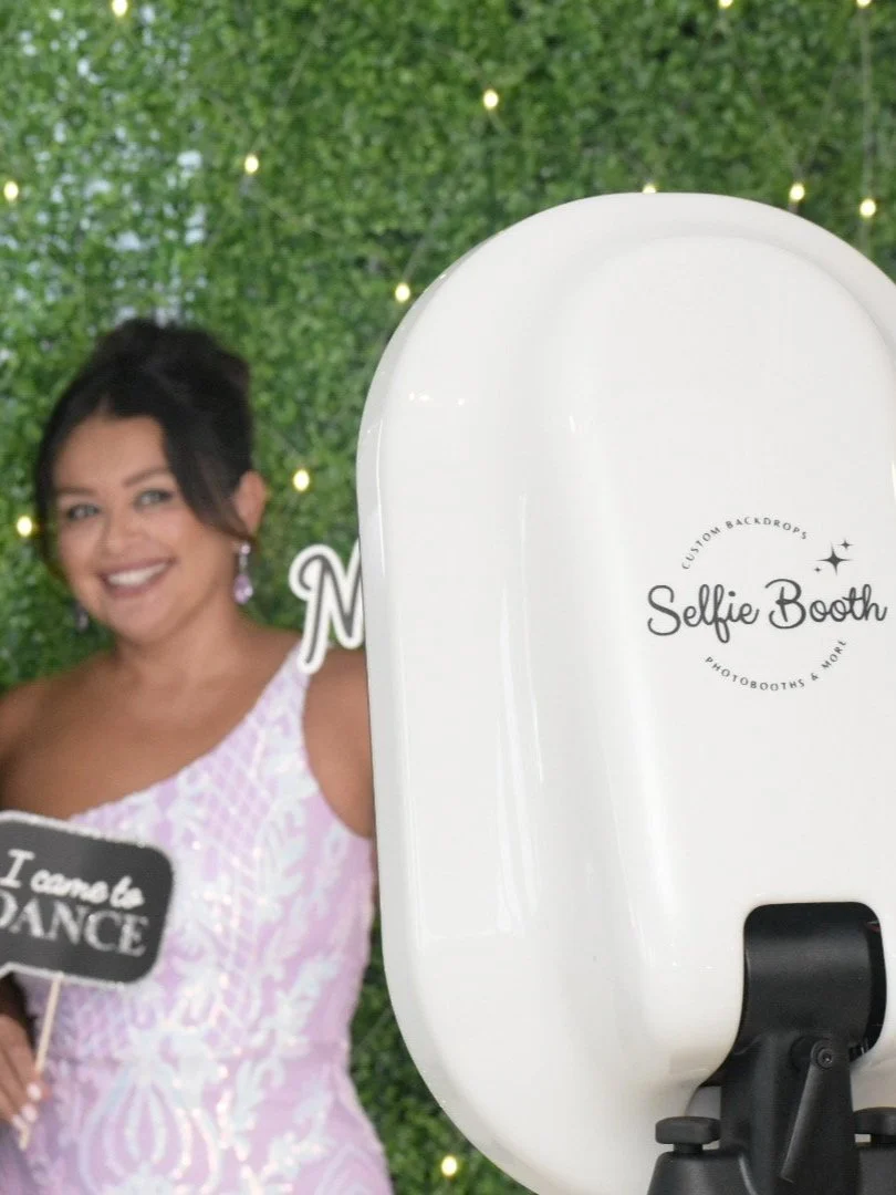 Woman smiling in front of a green leafy backdrop with string lights, standing behind a white photo booth with "Selfie Booth" printed on it, holding a sign that says "I come to DANCE".